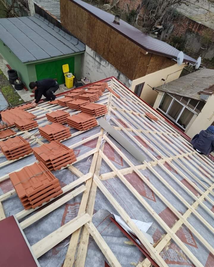 Montaż pomarańczowej dachówki na drewnianej konstrukcji dachu pokrytej membraną, widok z góry na budynki w tle.