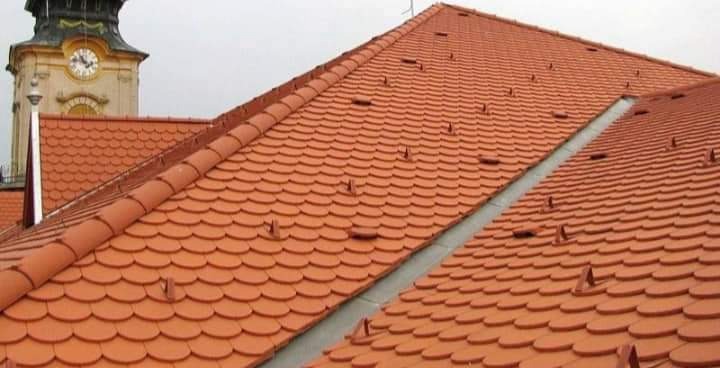 Dach pokryty czerwoną dachówką karpiówką z widocznymi zabezpieczeniami przeciwśniegowymi, w tle wieża zegarowa.