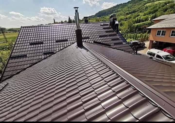 Nowy, brązowy dach z blachodachówki modułowej, z dwoma kominami, widok z góry na tle zielonych wzgórz i zabudowań.