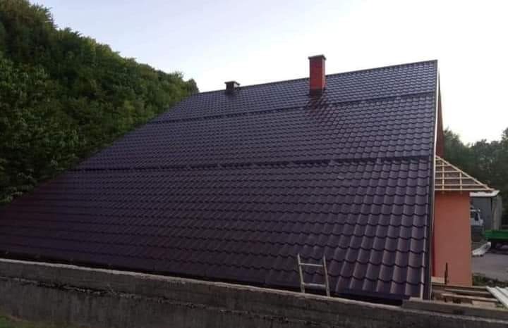 Nowy, ciemnobrązowy dach z blachodachówki z czerwonym kominem, widziany z dołu, z drabiną opartą o ścianę, a w tle drzewa i wznoszona konstrukcja dachu sąsiedniego budynku.