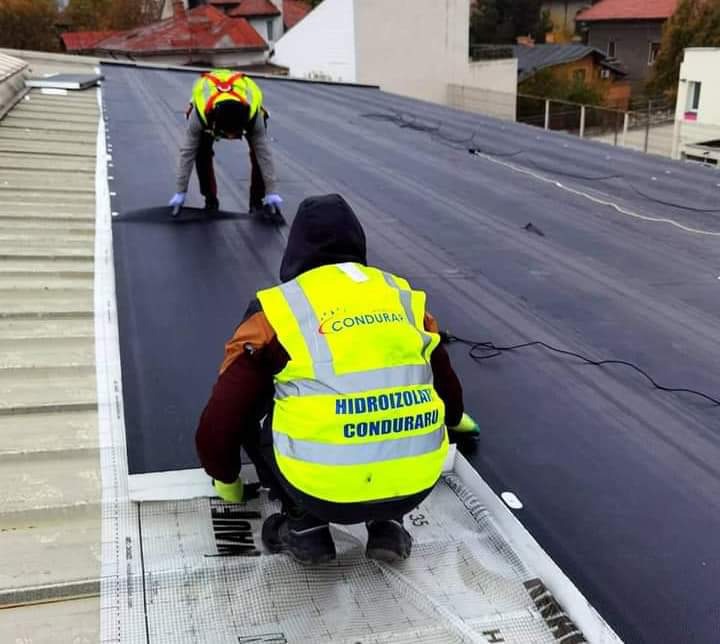 Dwóch pracowników w odblaskowych kamizelkach układa hydroizolację na płaskim dachu, widoczne rolki materiału i zgrzewarka.