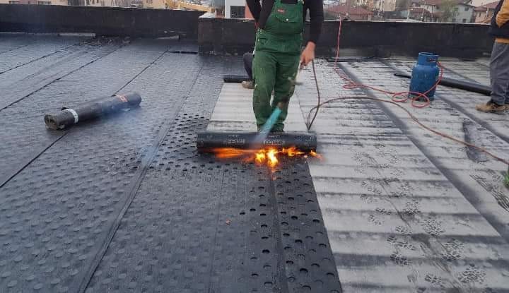 Pracownik w zielonym kombinezonie z palnikiem gazowym podczas układania papy termozgrzewalnej na dachu, widoczna butla gazowa i zgrzana powierzchnia dachu.