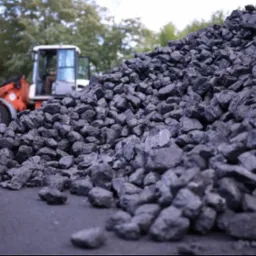 Góra czarnego węgla kamiennego na utwardzonym placu, w tle pomarańczowa ładowarka kołowa z operatorem w kabinie.