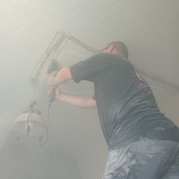 Elektric Serwis Krzysztof Baranowski - Elektryk w okularach, wiercący otwór w suficie pod instalację oświetleniową, widoczny kabel i lampa. Praca w trakcie wykonywania w zapylonym pomieszczeniu.