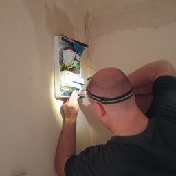 Elektric Serwis Krzysztof Baranowski - Elektryk z lampą czołową, w trakcie podłączania przewodów w skrzynce rozdzielczej na ścianie, w pomieszczeniu o surowym wykończeniu.