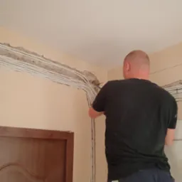 Elektryk montuje przewody w ścianie. Widoczne liczne kable w białych osłonach, prowadzone wzdłuż ściany pod sufitem. W tle fragment drzwi.