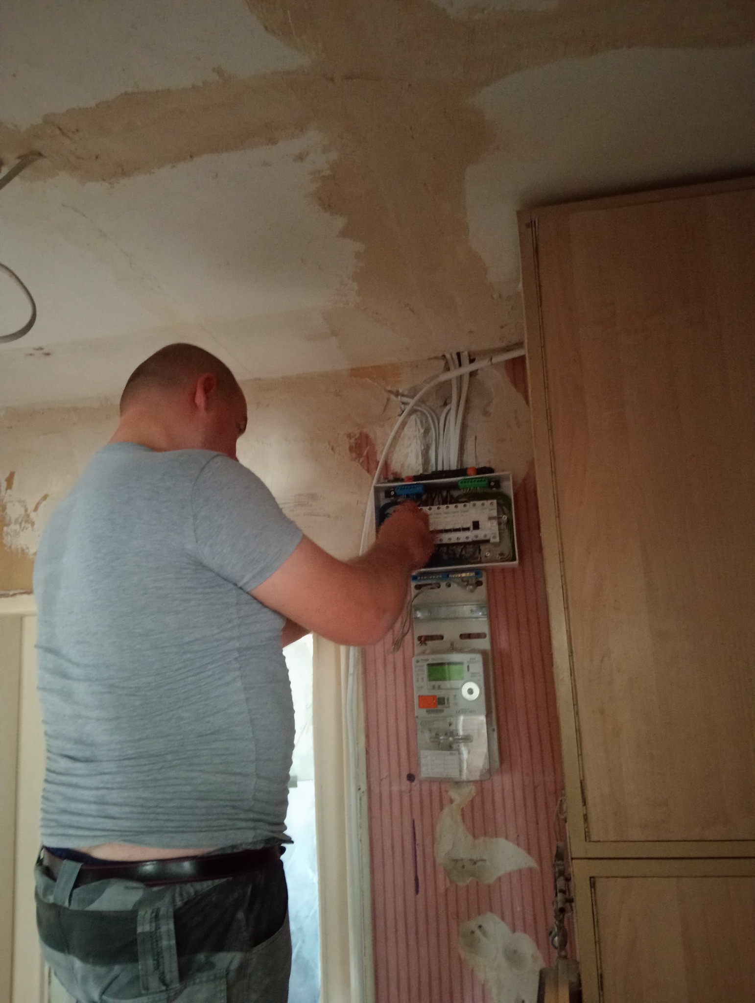 Elektryk w szarej koszulce pracuje nad instalacją elektryczną, widoczna skrzynka z bezpiecznikami i liczniki w pomieszczeniu w trakcie remontu.