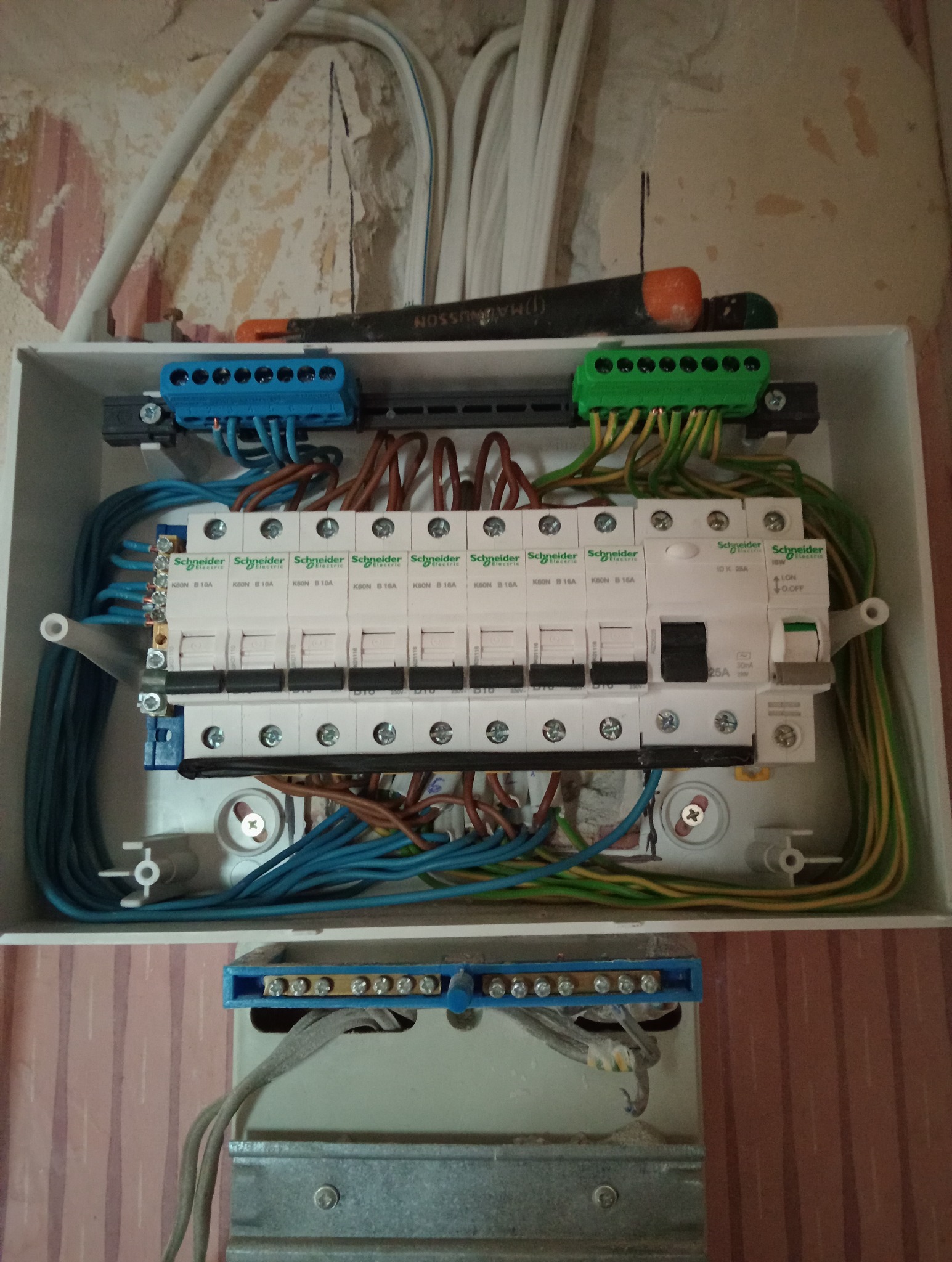 Rozdzielnica elektryczna z bezpiecznikami Schneider, okablowaniem w kolorach niebieskim, brązowym i zielono-żółtym. Nad rozdzielnicą leży śrubokręt.