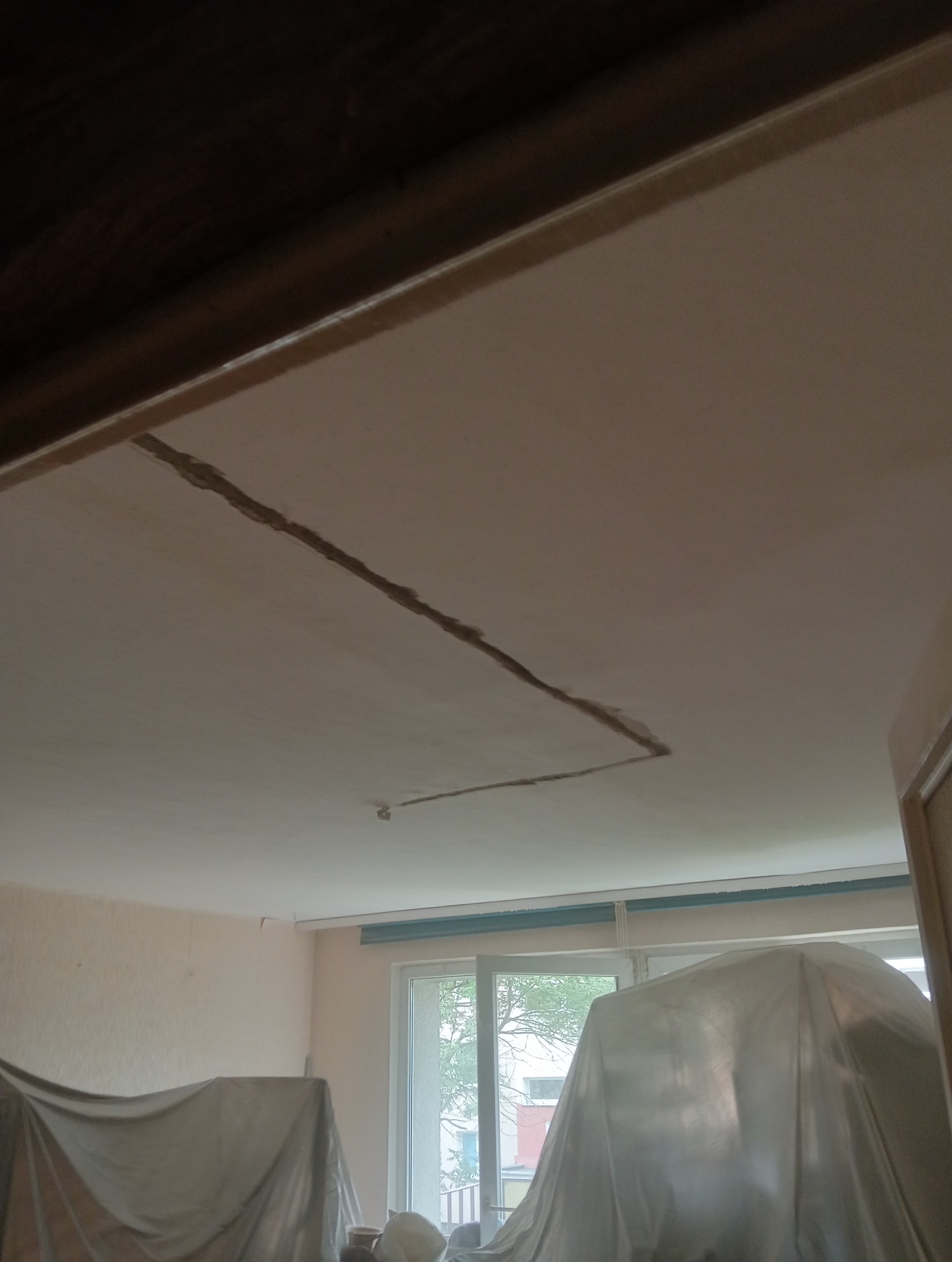 Biały sufit z widocznym pęknięciem przed naprawą. Pokój zabezpieczony folią malarską, okno widoczne w tle. Ujęcie z dołu, perspektywa z wnętrza pomieszczenia.