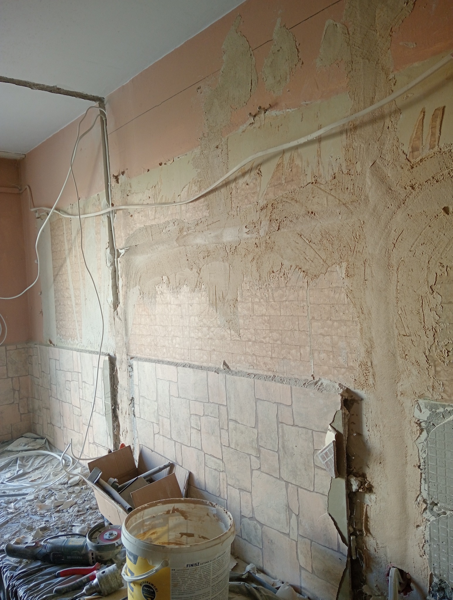 Remont pokoju: zerwane płytki, przygotowana ściana do gładzi, widoczne kable elektryczne,  narzędzia i wiadro z gładzią. Etap prac wykończeniowych.