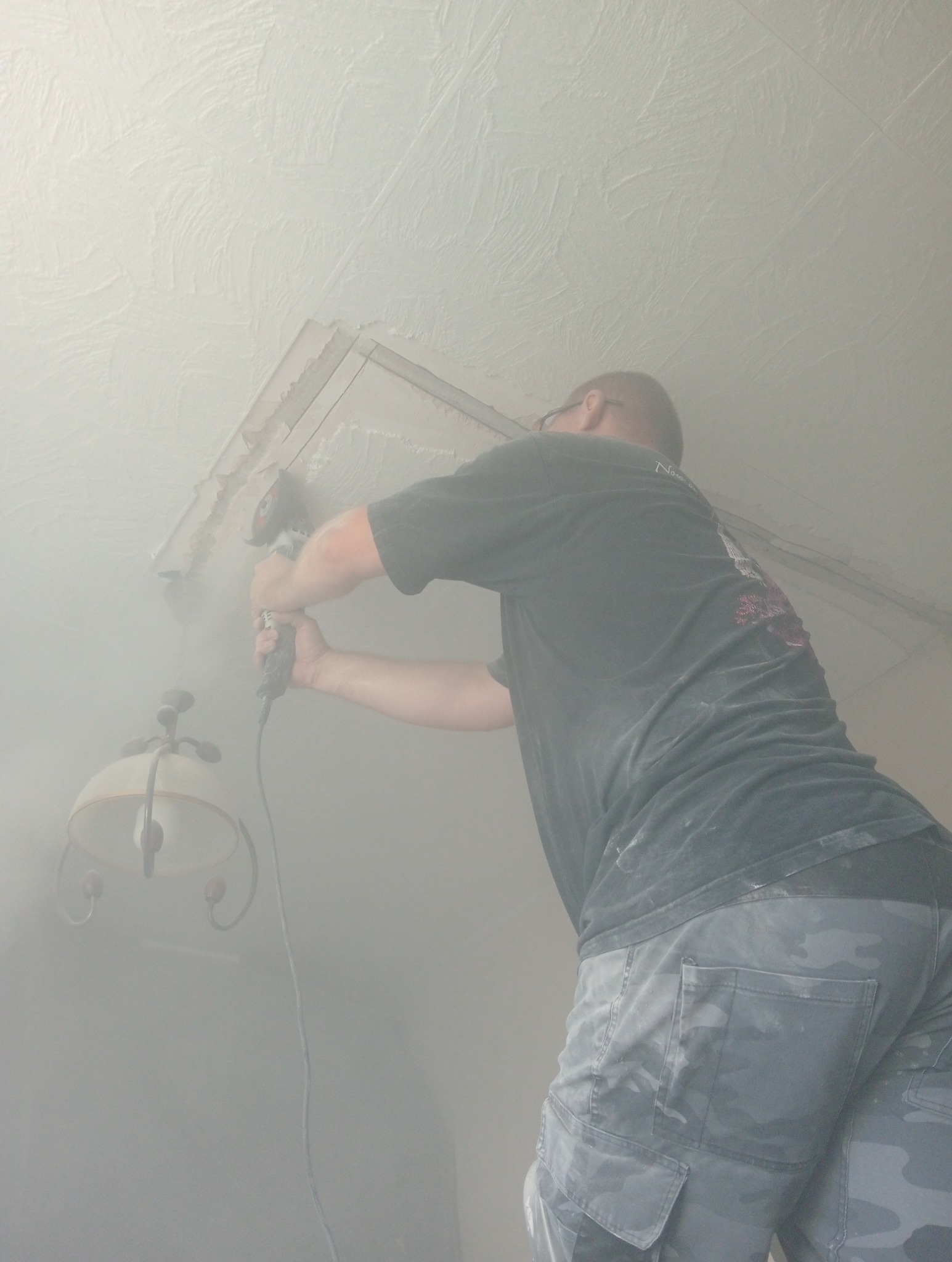 Elektryk w okularach, wiercący otwór w suficie pod instalację oświetleniową, widoczny kabel i lampa. Praca w trakcie wykonywania w zapylonym pomieszczeniu.