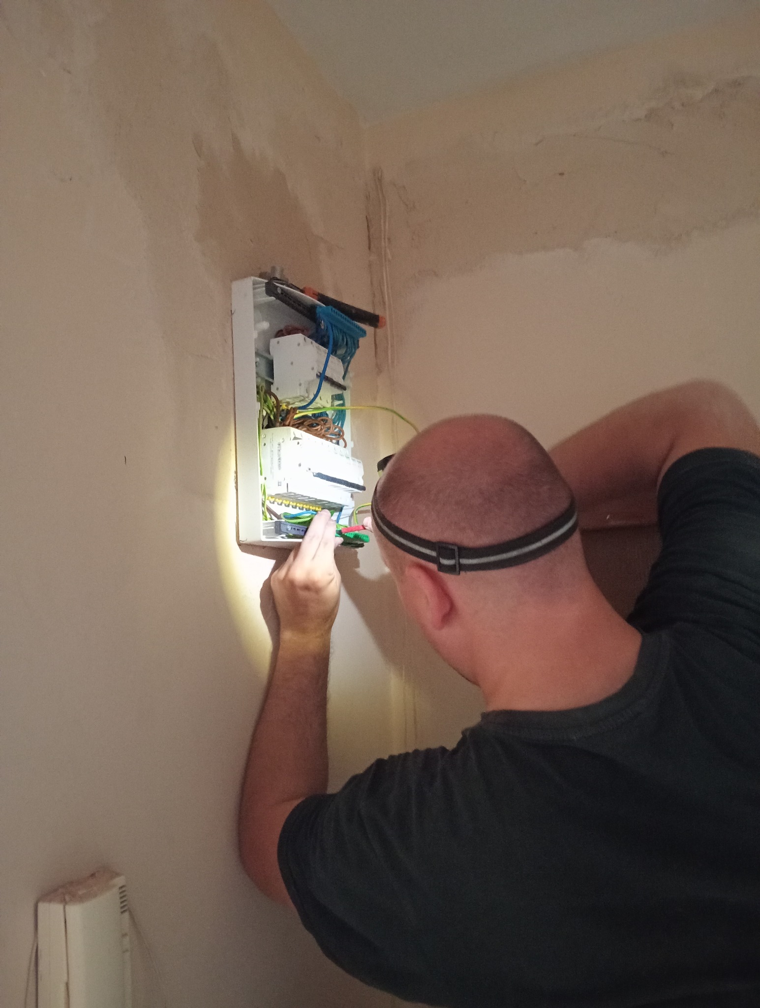 Elektryk z lampą czołową, w trakcie podłączania przewodów w skrzynce rozdzielczej na ścianie, w pomieszczeniu o surowym wykończeniu.