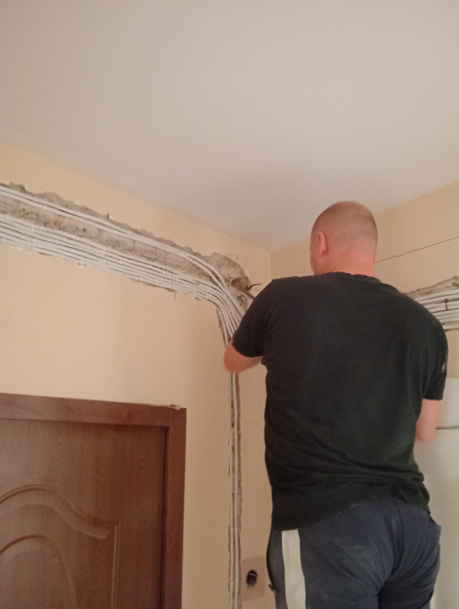 Elektryk montuje przewody w ścianie. Widoczne liczne kable w białych osłonach, prowadzone wzdłuż ściany pod sufitem. W tle fragment drzwi.