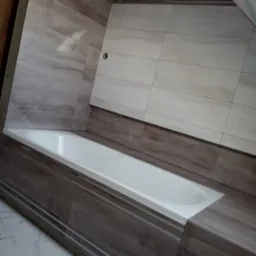 Biała wanna obudowana brązowymi płytkami imitującymi drewno, ściany wyłożone częściowo brązowymi i białymi płytkami, widoczne niedokończone prace przy suficie.