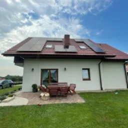 Dom jednorodzinny z panelami słonecznymi na dachu z czerwonej dachówki, widok z ogrodu, w tle błękitne niebo z chmurami.