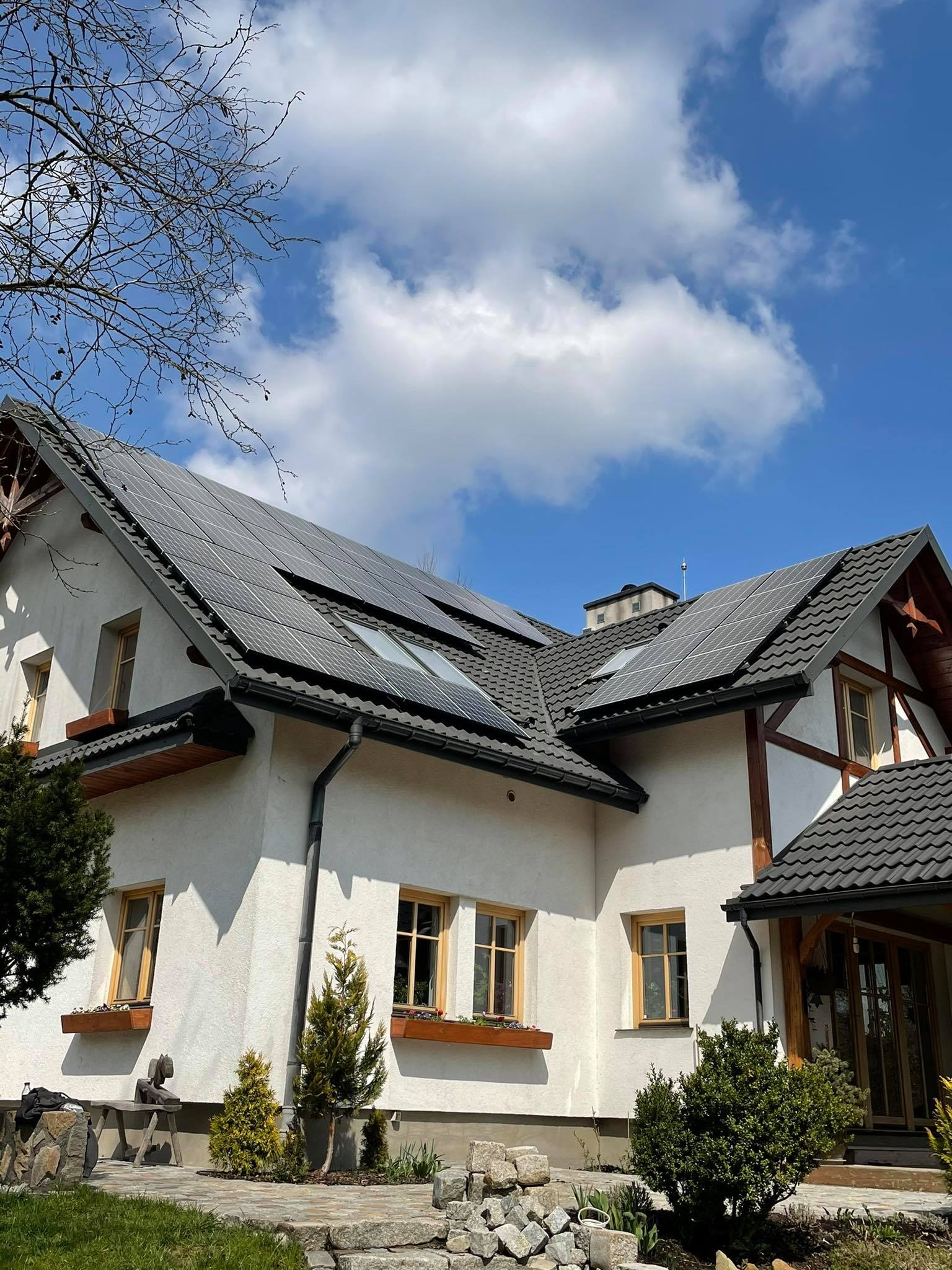 Biały dom z ciemną dachówką i panelami słonecznymi na dachu, widok z poziomu gruntu w słoneczny dzień z błękitnym niebem i chmurami.