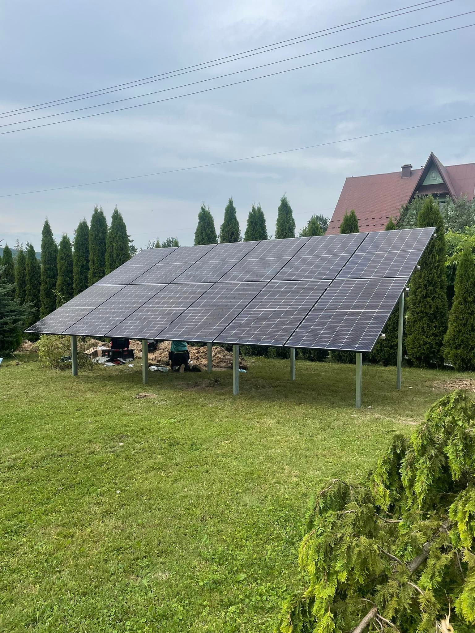 Panel słoneczny na metalowej konstrukcji w ogrodzie, widoczne narzędzia i osoba pracująca przy instalacji, dom z czerwonym dachem w tle.