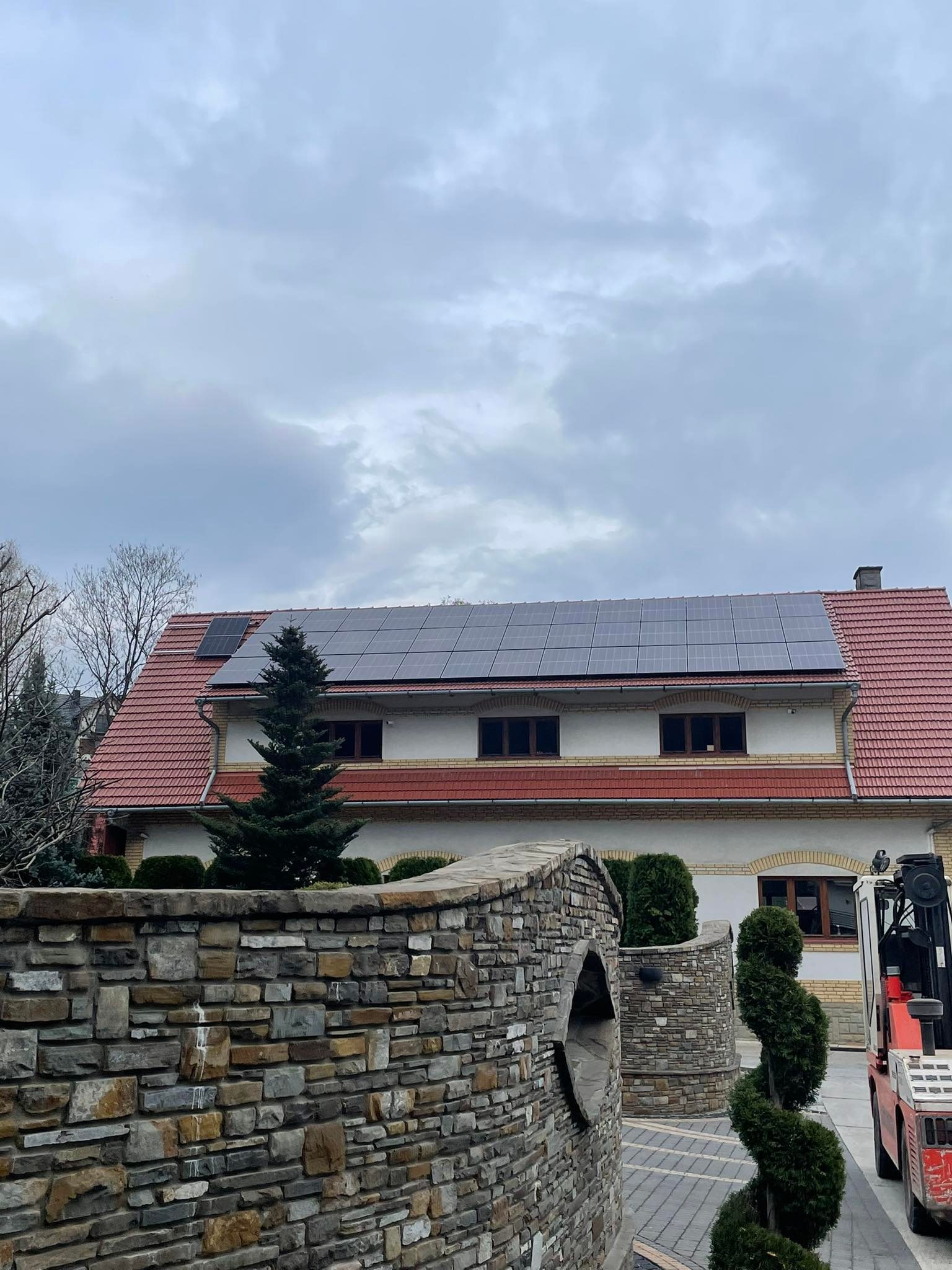 Dwukondygnacyjny dom z czerwoną dachówką i panelami słonecznymi na dachu, widoczny zza kamiennego muru i ozdobnych krzewów, z wózkiem widłowym zaparkowanym obok.