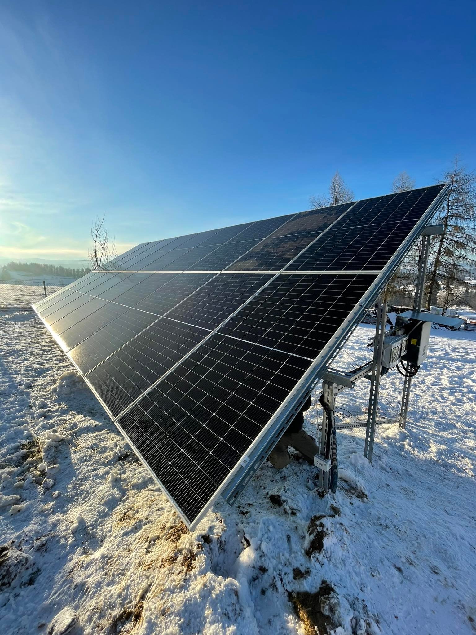 Panel fotowoltaiczny zamontowany na metalowej konstrukcji, pokryty śniegiem w zimowym krajobrazie, widoczne okablowanie.