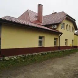 Żółta elewacja domu z brązowym pasem u dołu i czerwoną dachówką, widoczny komin i klimatyzator na ścianie.