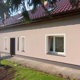 Jasny, jednopiętrowy dom z nową, jasnoróżową elewacją. Widoczne białe obramowania okien i drzwi, brązowy dach z dachówki i częściowo zagospodarowany teren przed budynkiem. Na drugim planie drabina...