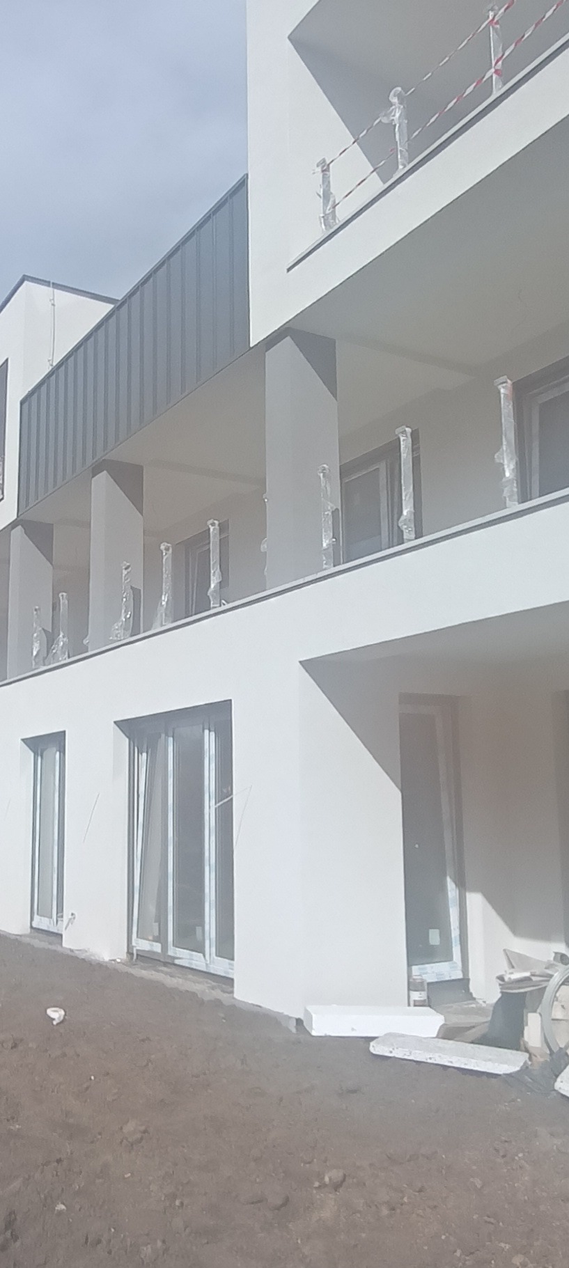 Nowoczesny budynek wielorodzinny z białą elewacją, szarym dachem i balkonami z barierkami zabezpieczonymi folią ochronną, teren wokół budynku w trakcie prac wykończeniowych.