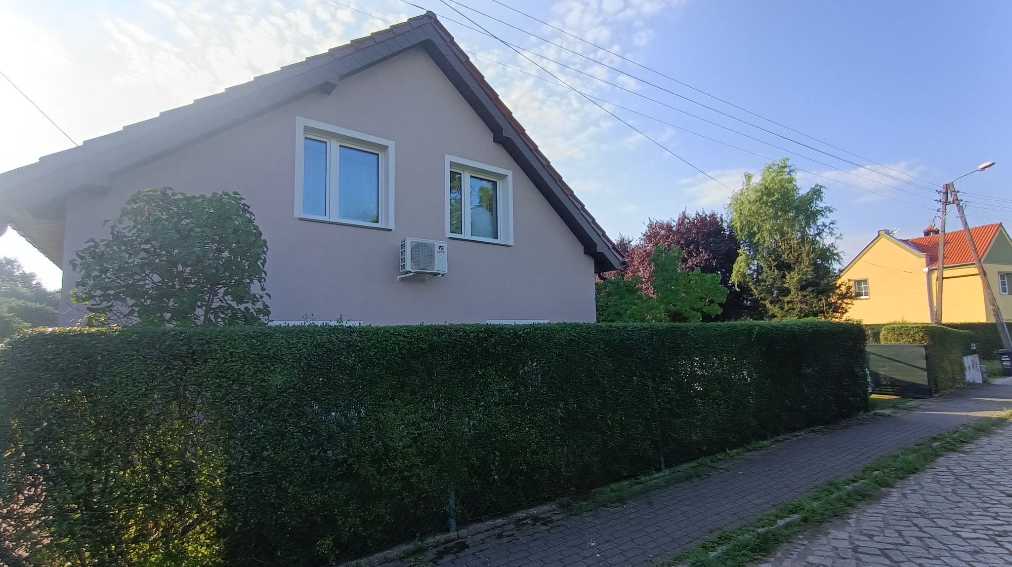 Jasnoróżowy dom jednorodzinny z dwoma oknami, klimatyzatorem zewnętrznym i ciemnobrązowym dachem, otoczony żywopłotem, widoczny z chodnika.