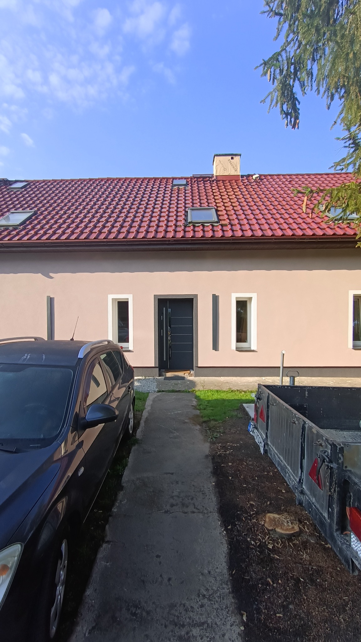 Dom jednorodzinny z nową, beżową elewacją i czerwoną dachówką, widoczne okna dachowe, zaparkowany samochód i przyczepa.