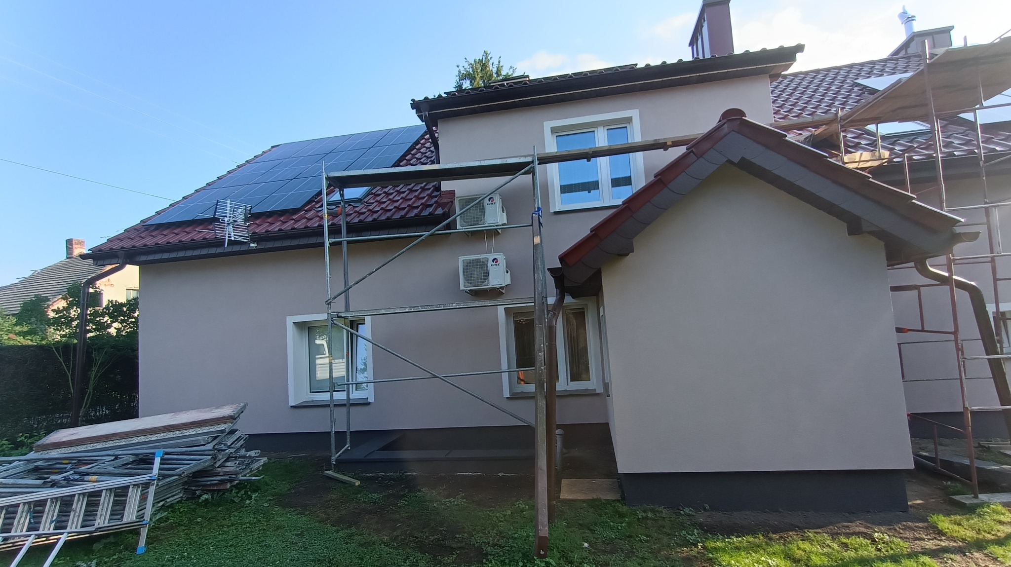Dom jednorodzinny z nową, jasnoszarą elewacją, widoczne rusztowania, panele słoneczne na dachu i klimatyzatory na ścianie budynku.