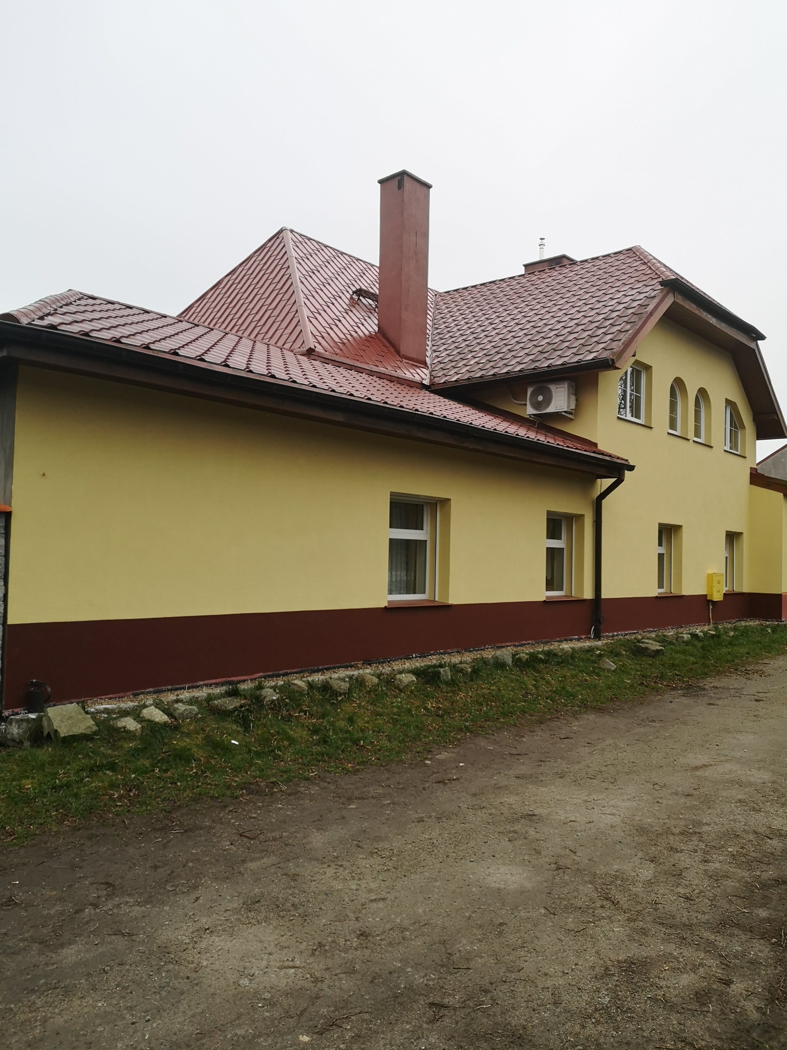 Żółta elewacja domu z brązowym pasem u dołu i czerwoną dachówką, widoczny komin i klimatyzator na ścianie.