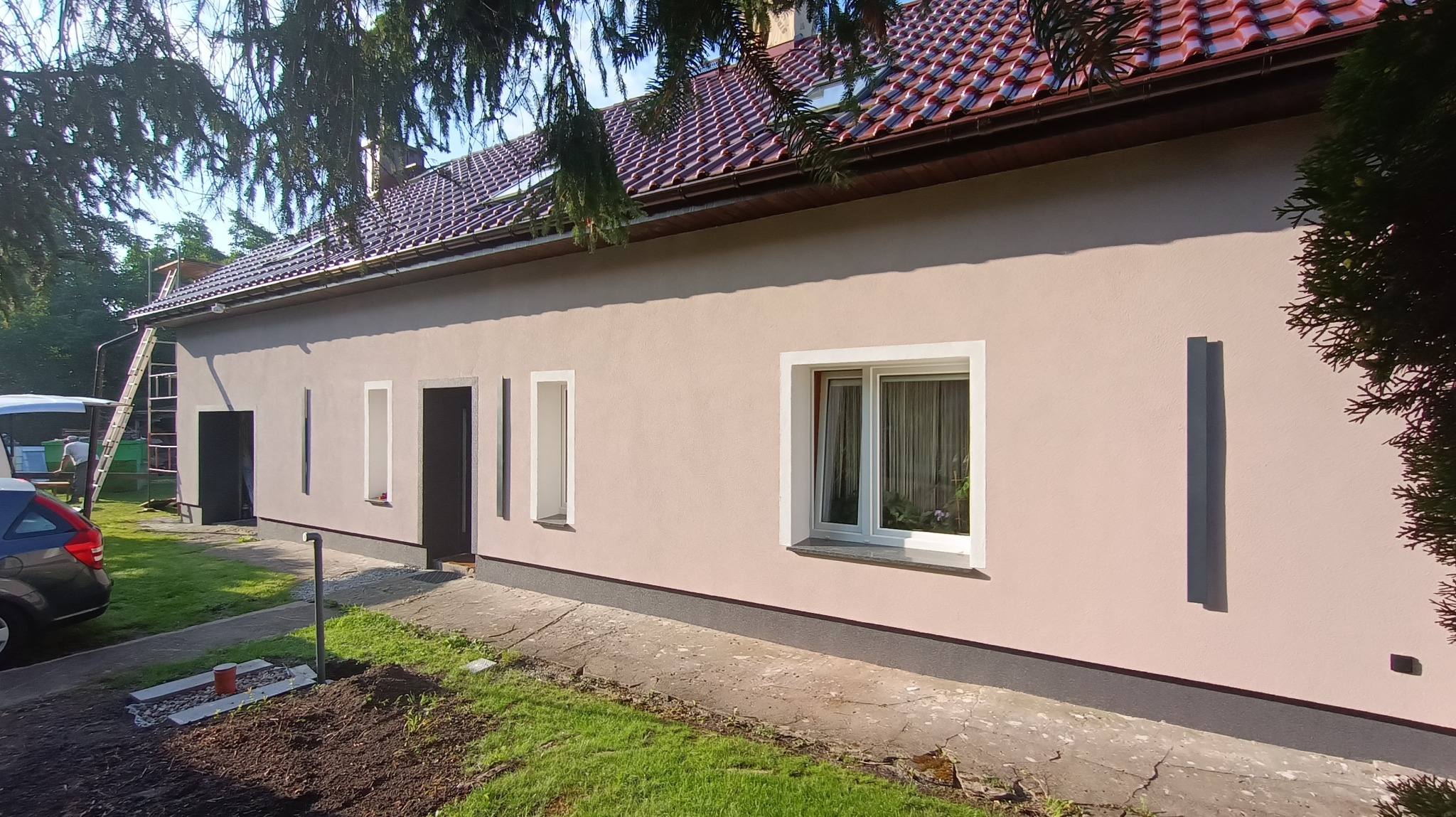 Jasny, jednopiętrowy dom z nową, jasnoróżową elewacją. Widoczne białe obramowania okien i drzwi, brązowy dach z dachówki i częściowo zagospodarowany teren przed budynkiem. Na drugim planie drabina...