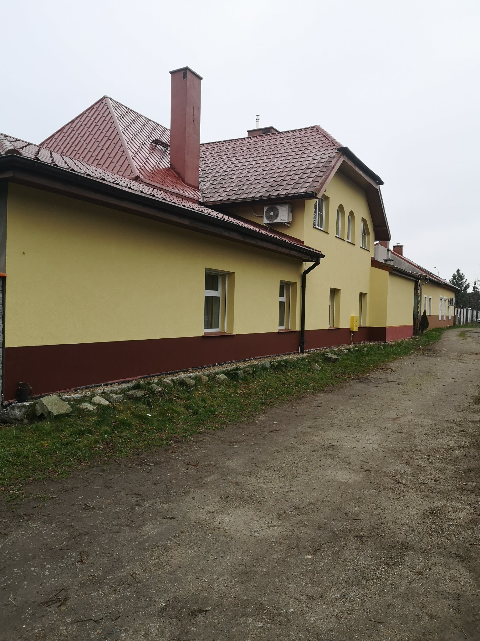 Żółty budynek z czerwoną dachówką, widoczny z boku, z elewacją w dwóch odcieniach żółci i brązu, z klimatyzatorem na ścianie i chodnikiem z boku.