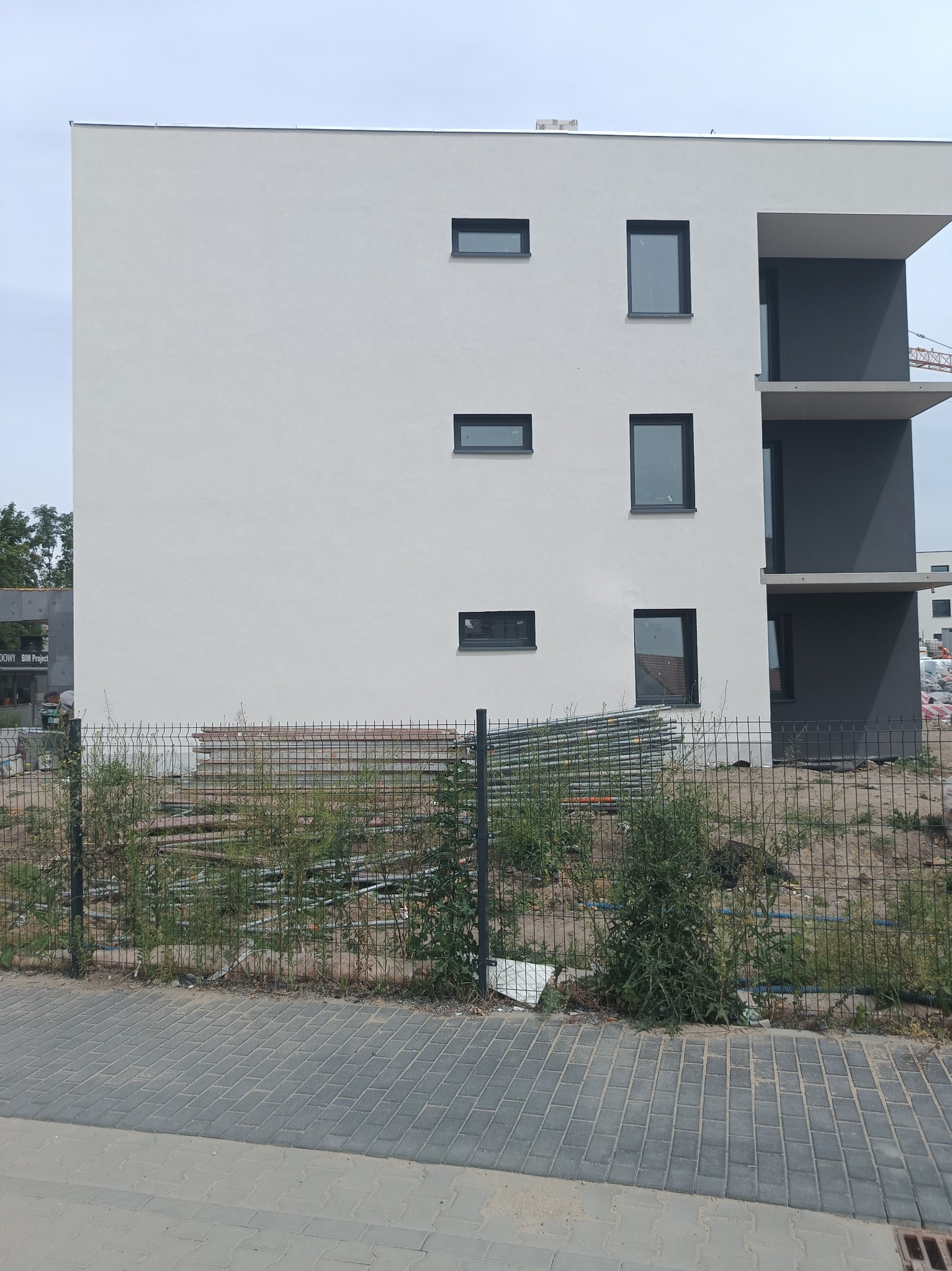 Nowoczesny budynek z jasnoszarą elewacją, czarnymi obramowaniami okien i balkonami w szarym odcieniu, widoczny przez metalowe ogrodzenie z elementami zieleni i stertą rur na placu budowy.