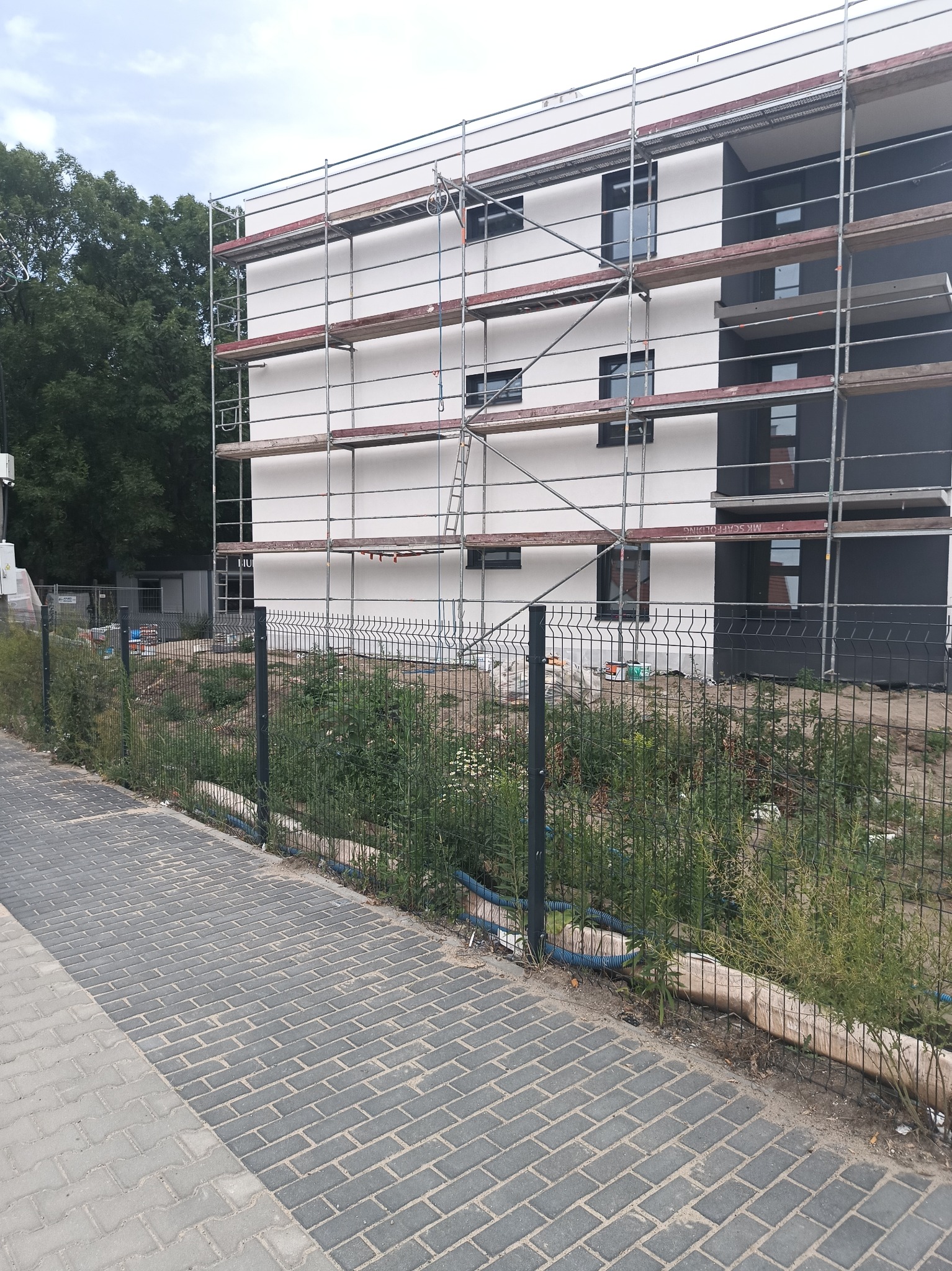 Nowoczesny budynek w trakcie prac elewacyjnych, z widocznymi rusztowaniami i fragmentami pomalowanej fasady w kolorze białym i grafitowym, ogrodzony siatką, z zielenią w tle.