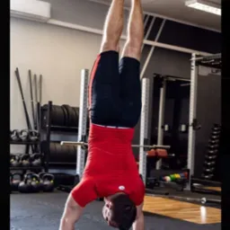 Mężczyzna w czerwonej koszulce i czarnych spodenkach wykonuje st&oacute;jkę na rękach w siłowni, w tle widoczne kettlebells i stojak na ciężary.