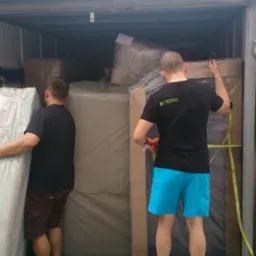 Dwóch mężczyzn w trakcie załadunku mebli do wnętrza ciężarówki, zabezpieczając je pasami transportowymi. Widoczny bok niebieskiej plandeki i biały van.