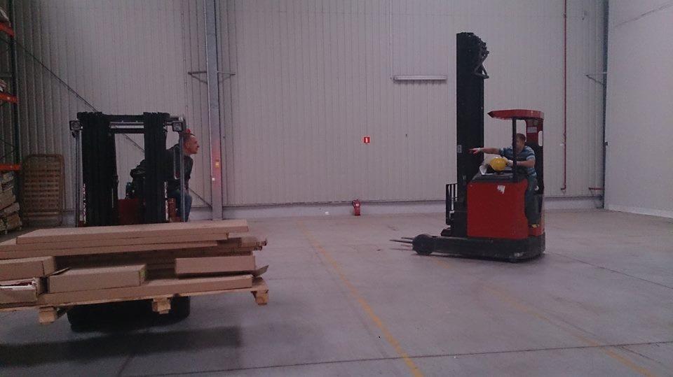 Dwa wózki widłowe w magazynie, jeden z paletą kartonów, drugi z operatorem wskazującym kierunek. Hala magazynowa z betonową podłogą i białymi ścianami.