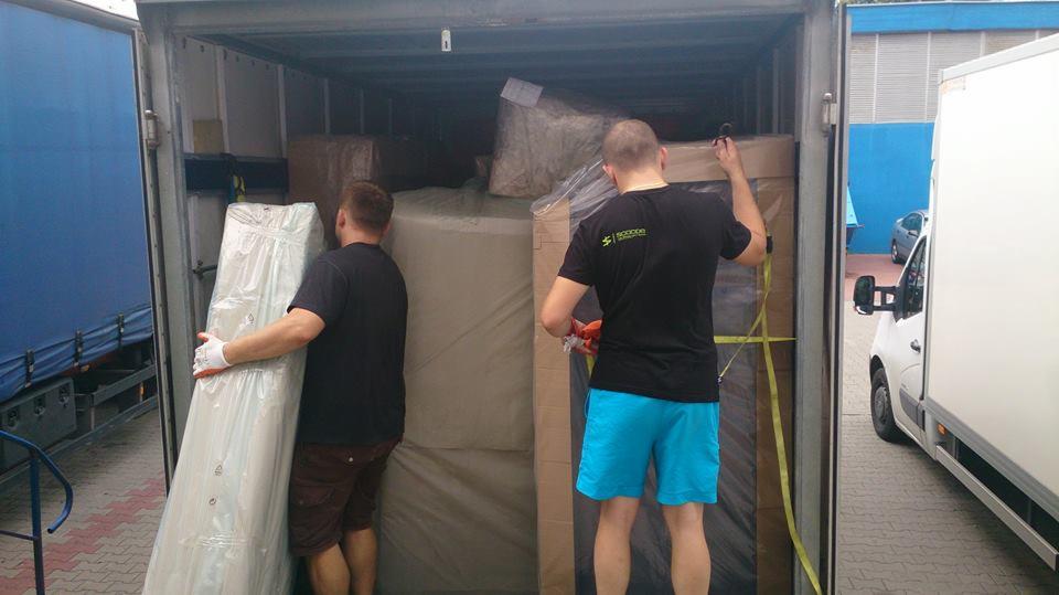 Dwóch mężczyzn w trakcie załadunku mebli do wnętrza ciężarówki, zabezpieczając je pasami transportowymi. Widoczny bok niebieskiej plandeki i biały van.