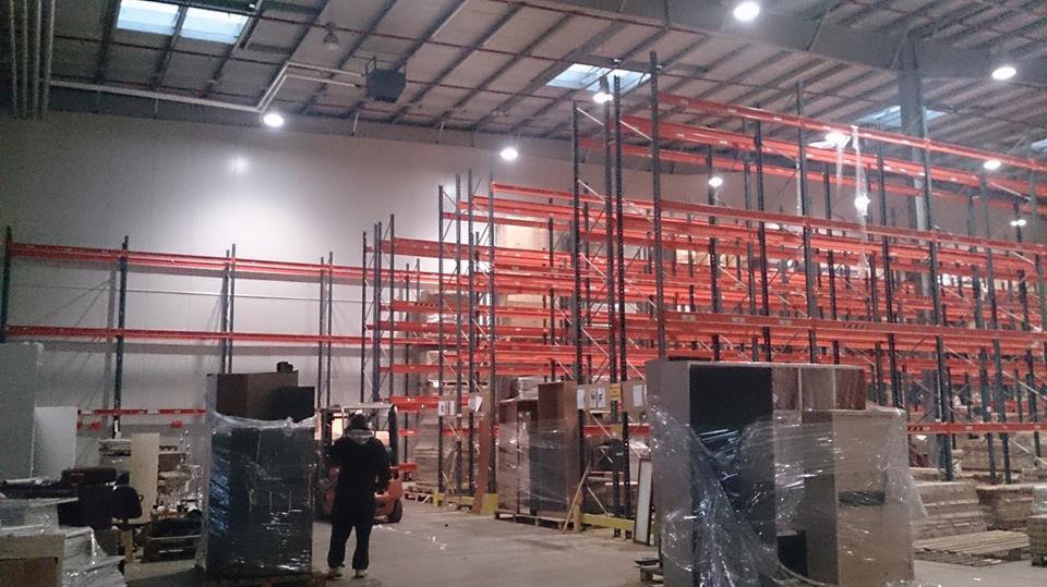 Magazyn z regałami, wózek widłowy i zapakowane kartony. Przygotowania do transportu, widoczny pracownik. Hala z oświetleniem sufitowym.