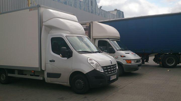 Dwa białe busy dostawcze Renault i Iveco, zaparkowane obok niebieskiej naczepy na tle szarego budynku. Renault Master z przodu, widoczna tablica rejestracyjna.