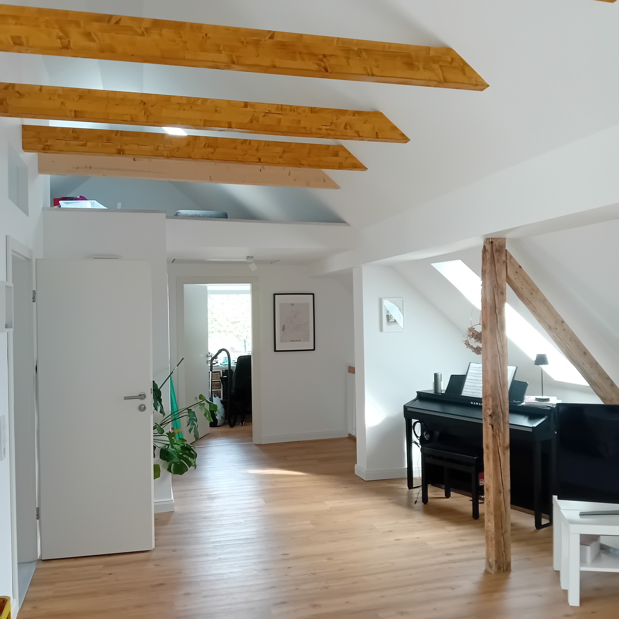 Jasne wnętrze poddasza z drewnianymi belkami stropowymi, pianinem i parkietem. Widok na otwarte drzwi i dalszą część mieszkania. Minimalistyczny design.