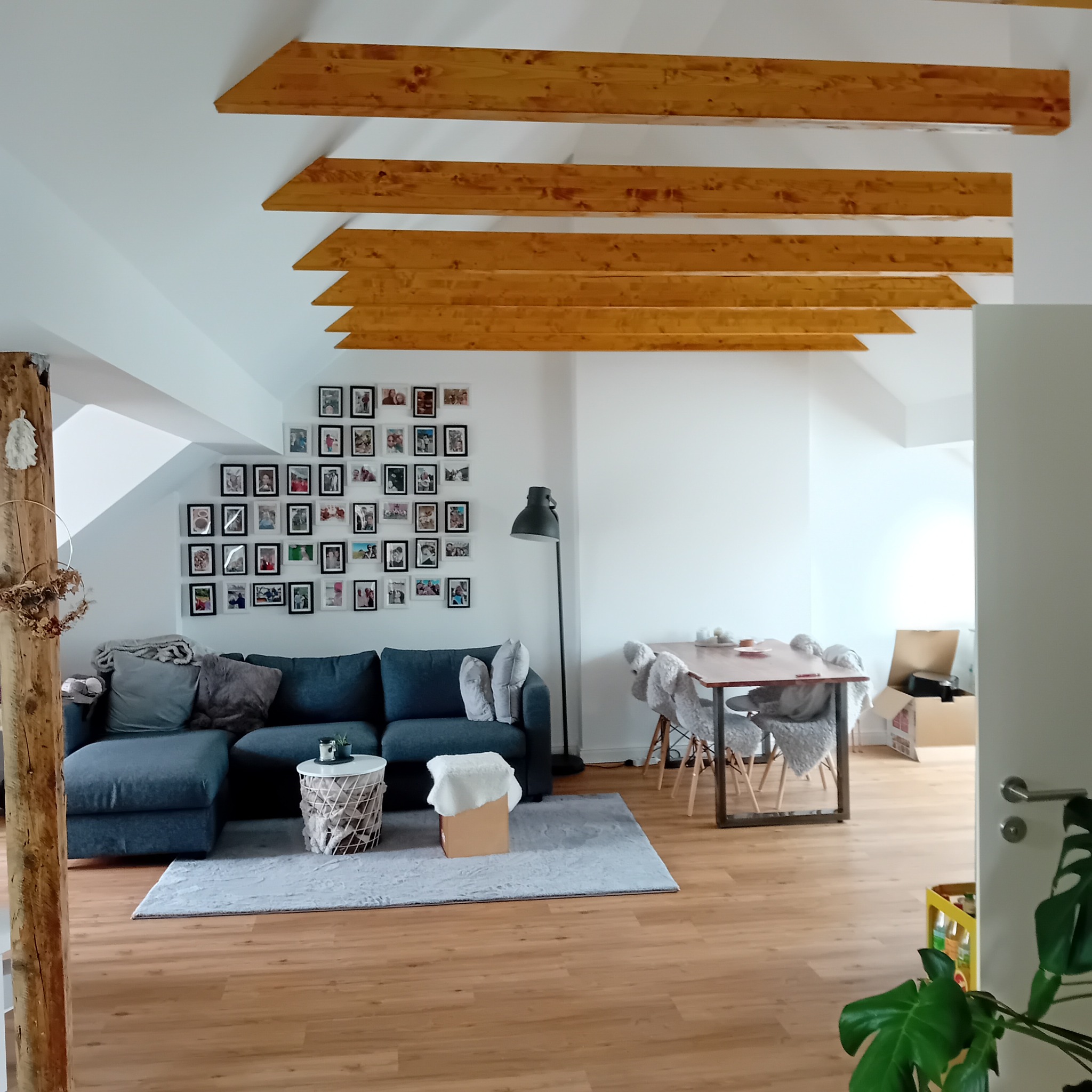 Jasne wnętrze poddasza z drewnianymi belkami stropowymi, szarą sofą i ścianą galerii zdjęć. Parkiet na podłodze. Widok na jadalnię z kartonami w tle.