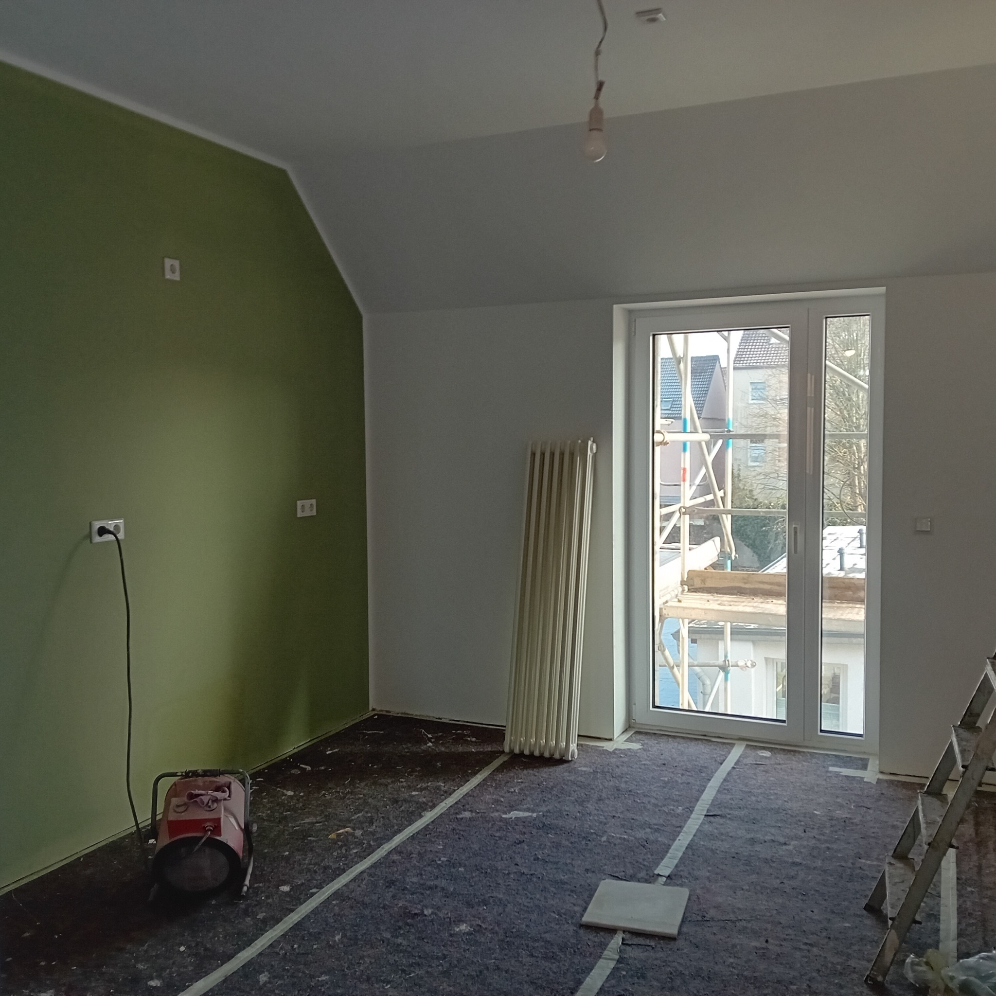 Pokój w trakcie malowania: zielona ściana, zabezpieczona podłoga, grzejnik i drabina. Widok z okna na rusztowanie.