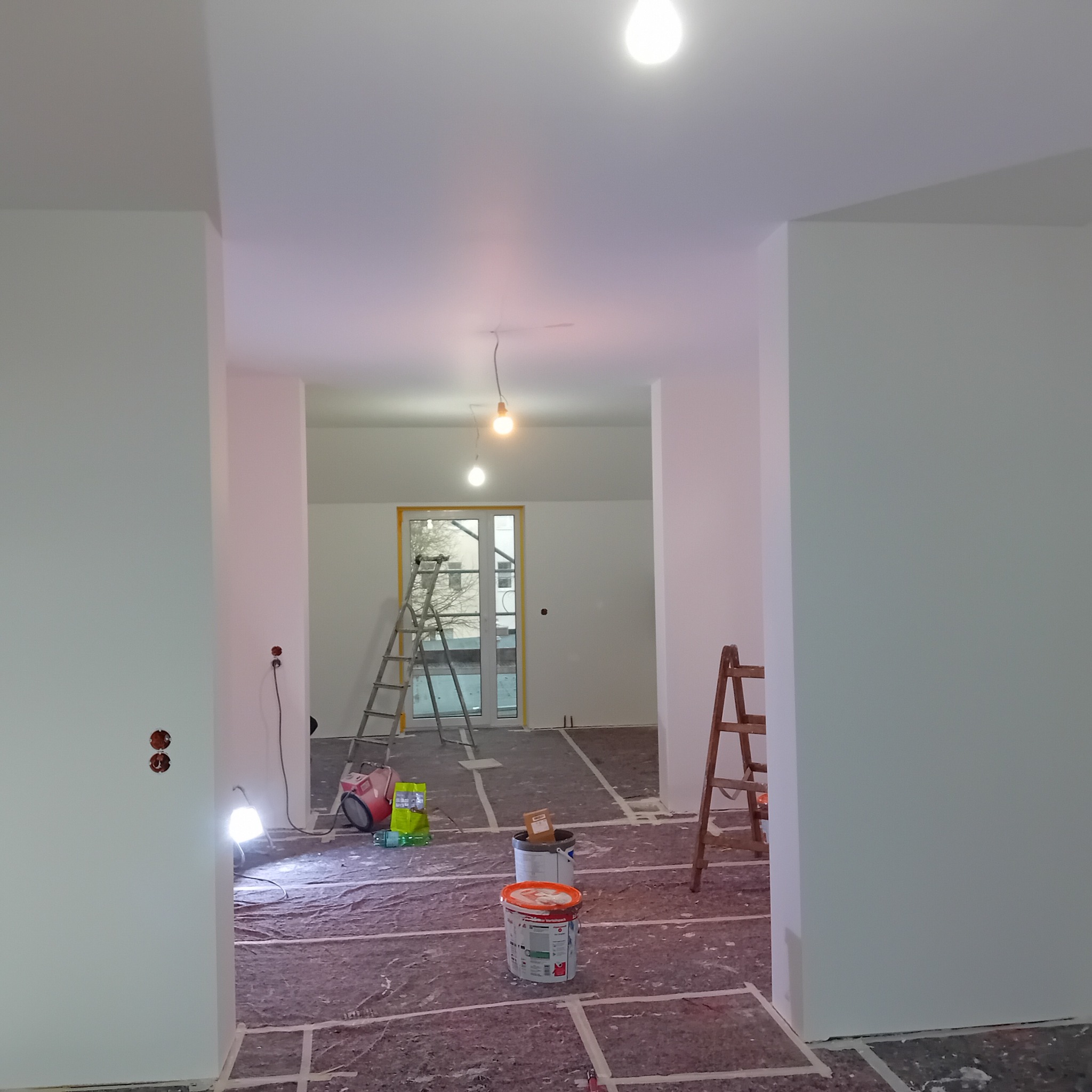 Wnętrze w trakcie malowania: białe ściany, zabezpieczona podłoga, drabiny, puszki z farbą i lampa halogenowa. Widok na okno z żółtą taśmą zabezpieczającą.