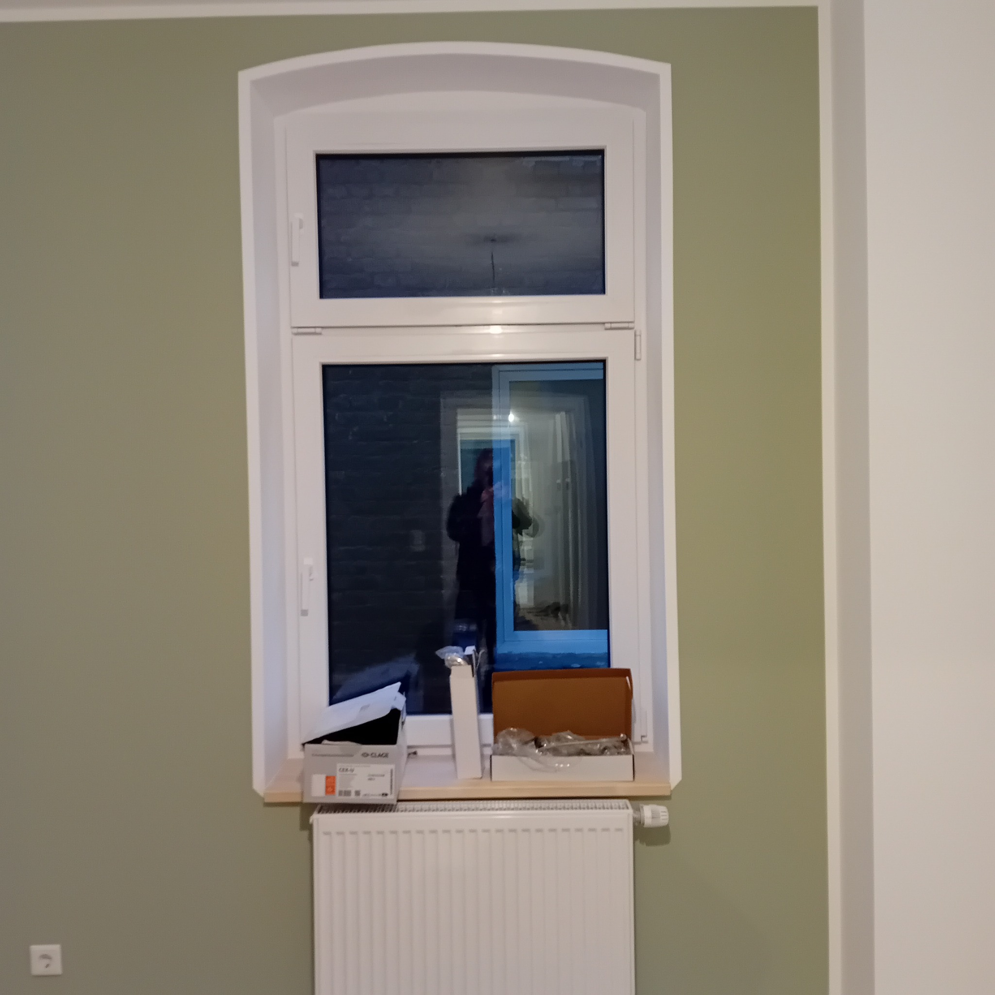 Białe okno PCV z zaokrąglonym górnym profilem, osadzone w ścianie pomalowanej na oliwkowy kolor. Na parapecie widoczne akcesoria montażowe i grzejnik.