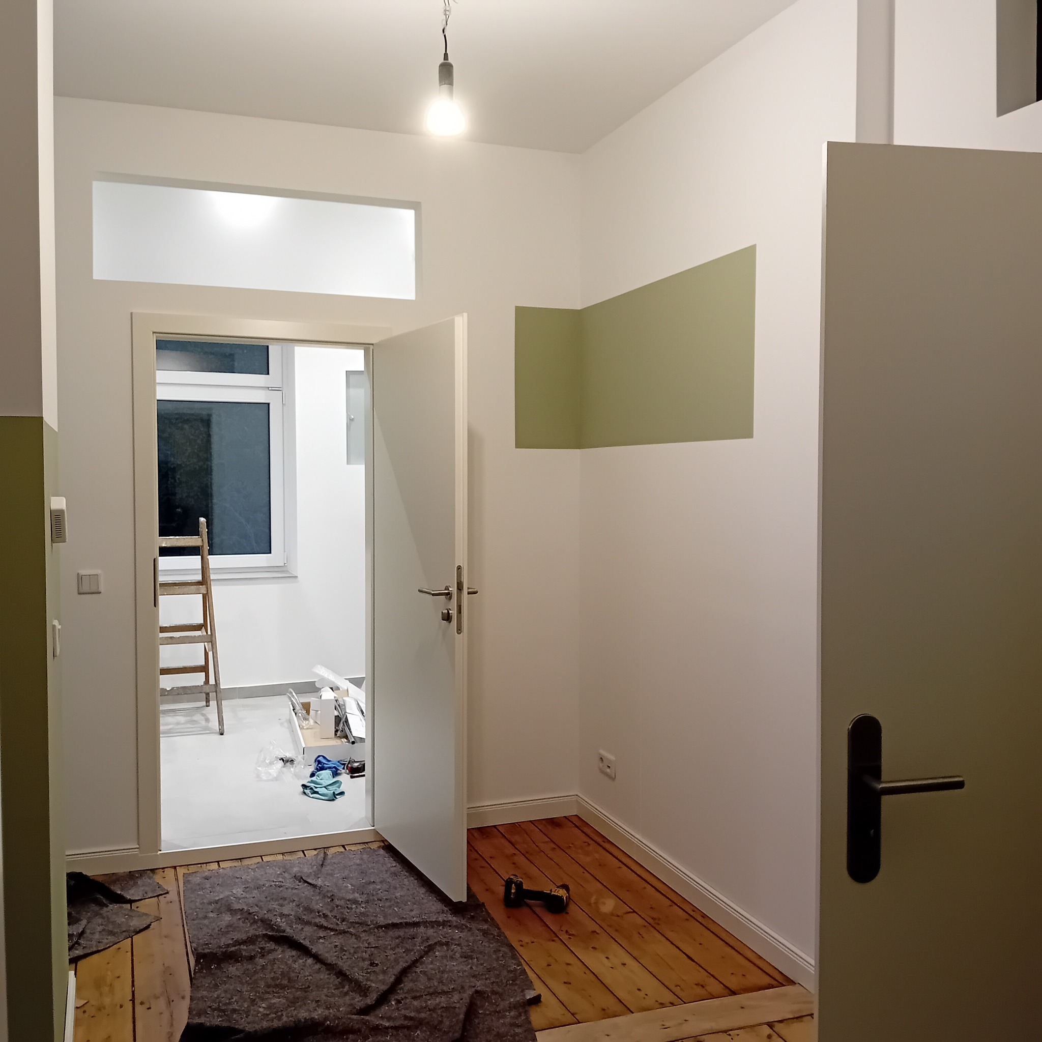 Wnętrze po częściowym malowaniu ścian na biało i oliwkowo, widoczne drewniane podłogi, drabina w sąsiednim pokoju i narzędzia malarskie. Otwarta przestrzeń, surowy wystrój.