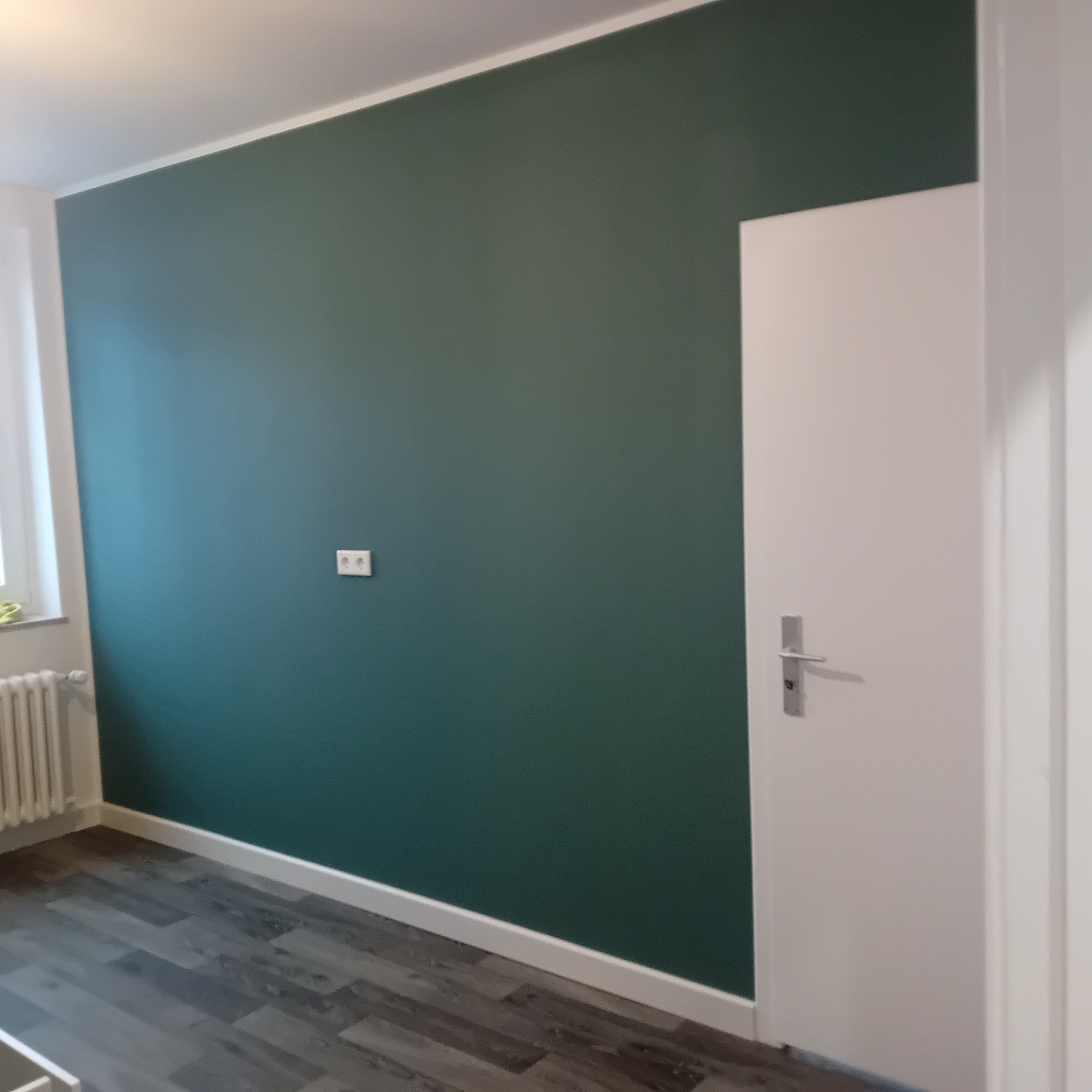 Pomieszczenie po malowaniu: ściana w odcieniu butelkowej zieleni, białe listwy przypodłogowe i sufitowe, szara podłoga imitująca drewno, białe drzwi z srebrną klamką.