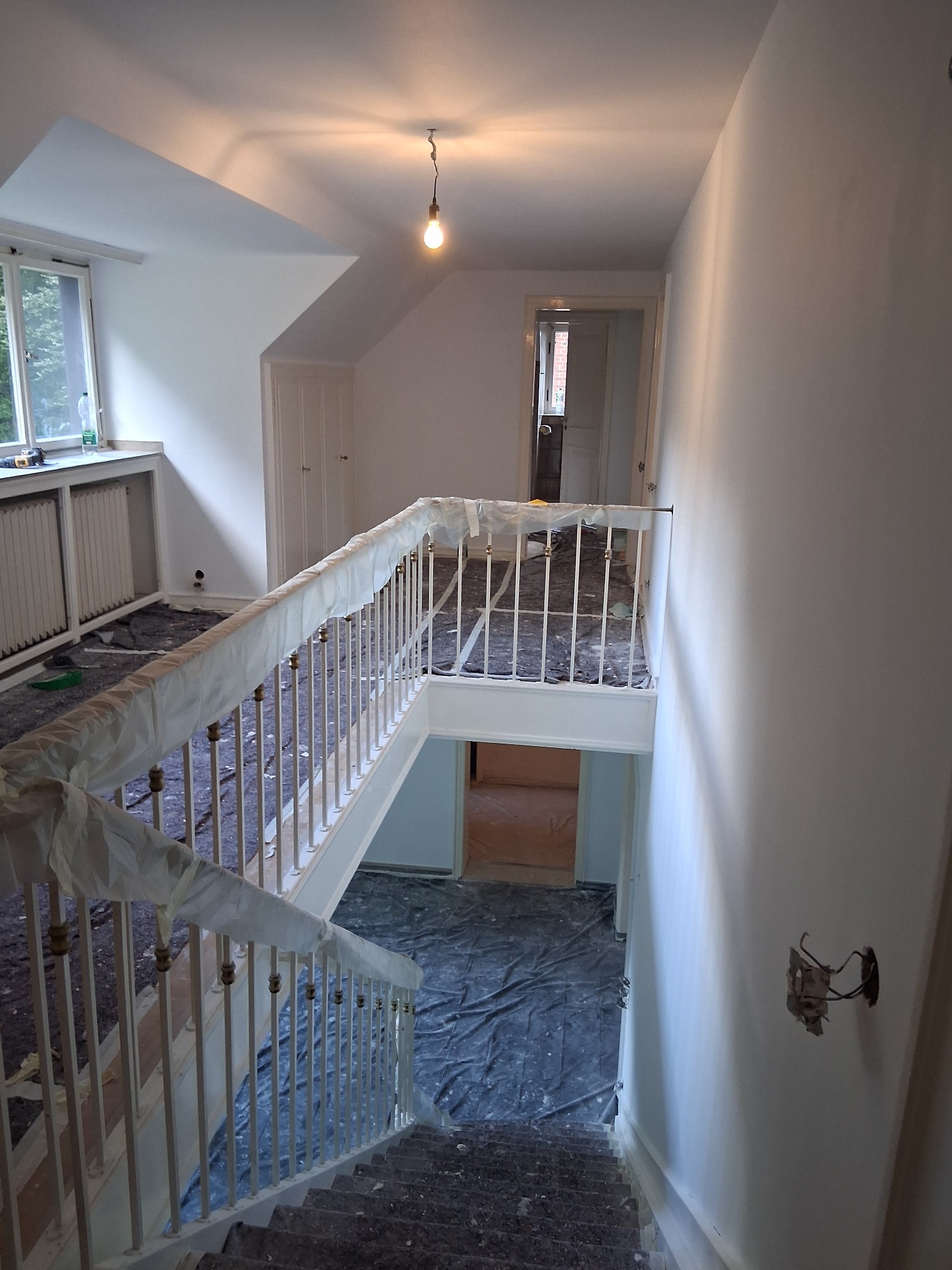 Klatka schodowa w trakcie malowania: zabezpieczone folią schody i balustrady, świeżo pomalowane ściany, widoczne gniazdko elektryczne bez osłony. Przygotowania do malowania.