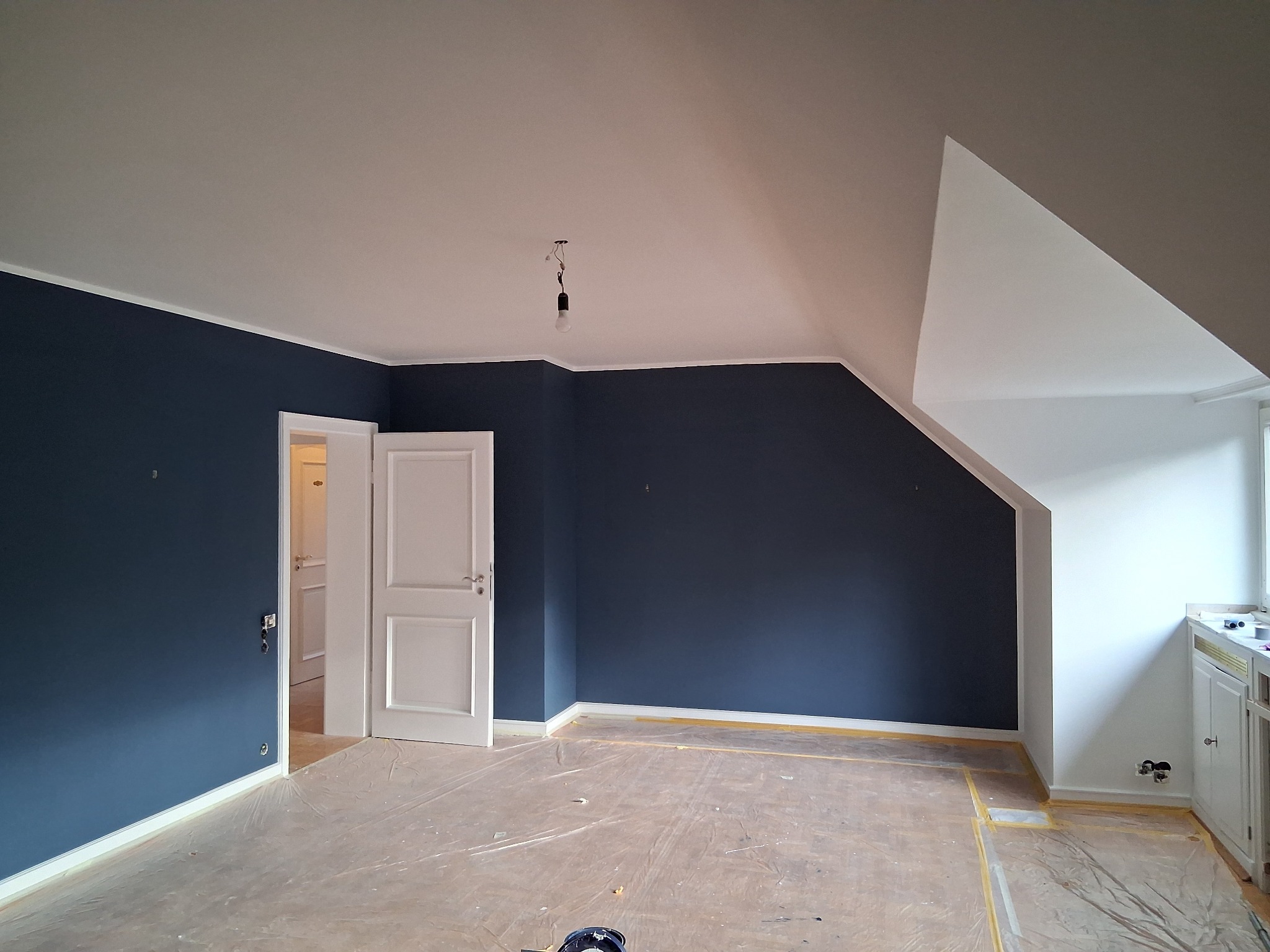 Pomalowane na granatowo ściany w pokoju na poddaszu z białym sufitem i listwami przypodłogowymi. Podłoga zabezpieczona folią malarską. Otwarta biała stolarka drzwiowa.