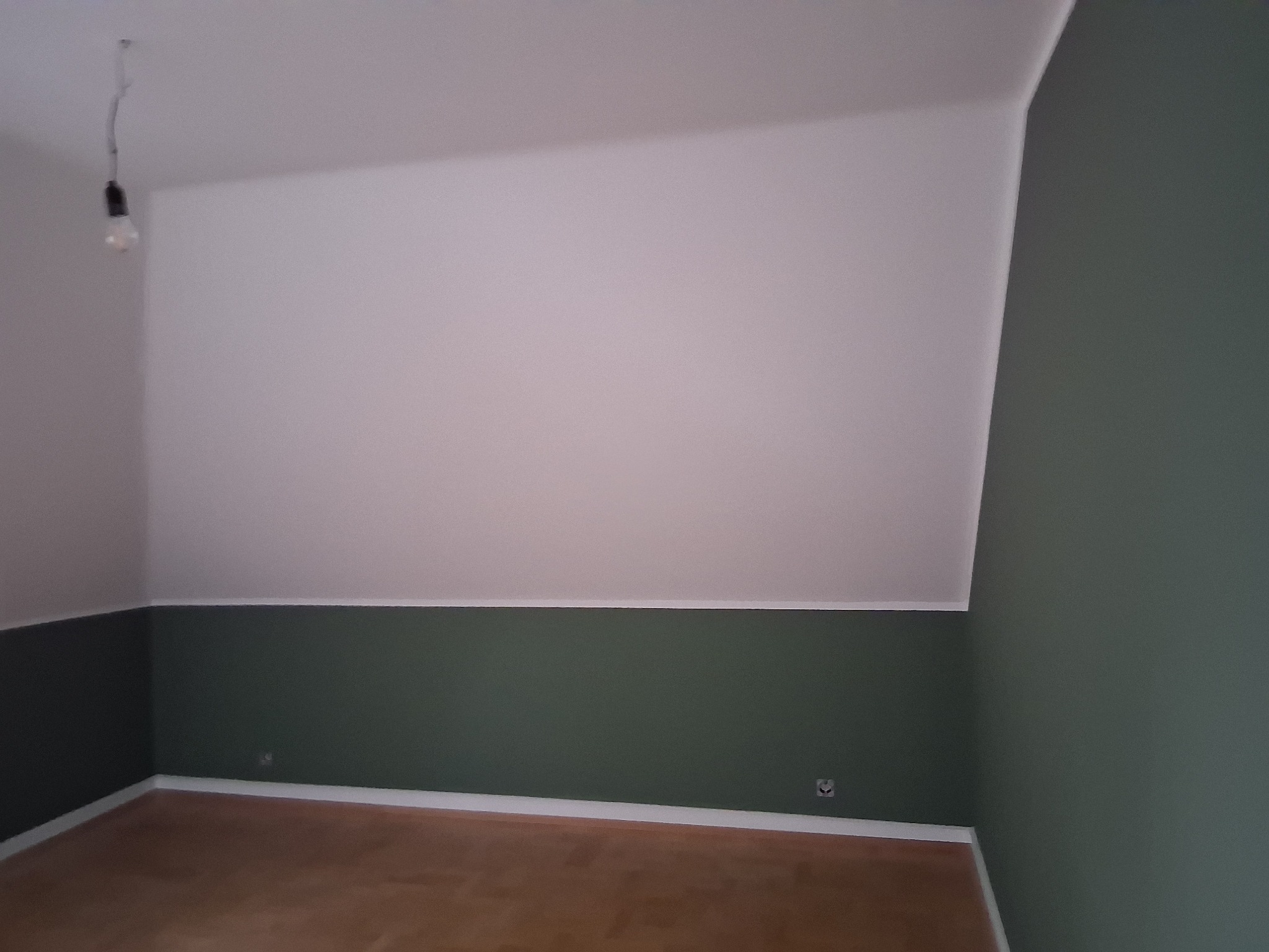 Pomieszczenie po malowaniu: biała góra, dół w odcieniach zieleni, drewniana podłoga. Widoczna żarówka na kablu. Świeżo pomalowane ściany w pokoju na poddaszu.